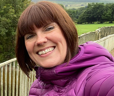 Bev Chapman outdoors in a purple jacket outdoors against a background of trees, smiling at the camera.