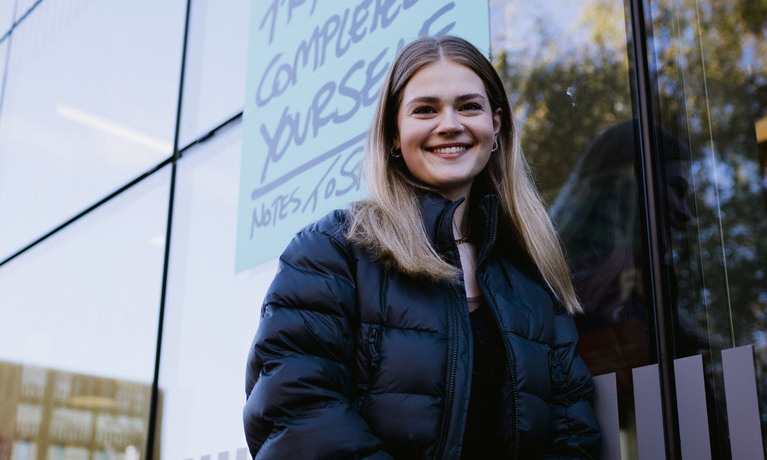 A student smiling against the large window wall of The Hub on campus.