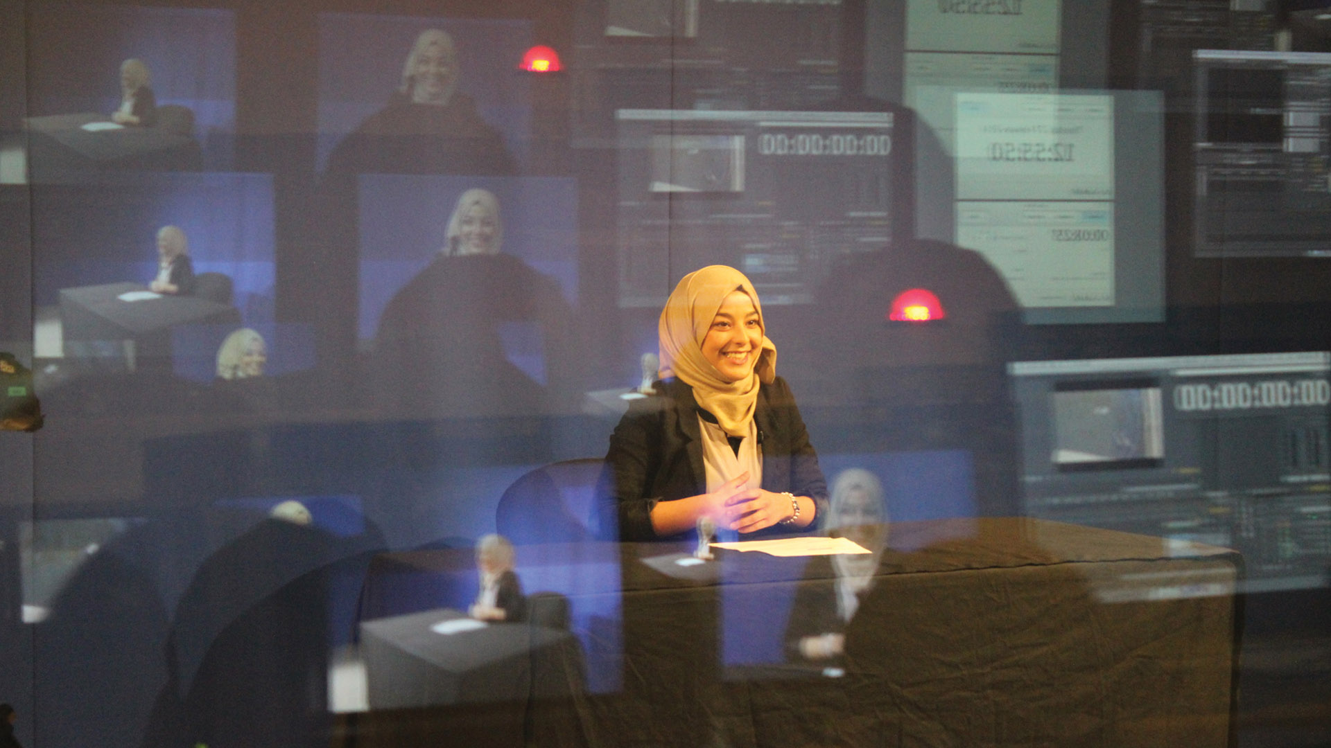 Smiling student stood in front of a TV screen wearing a yellow hijab