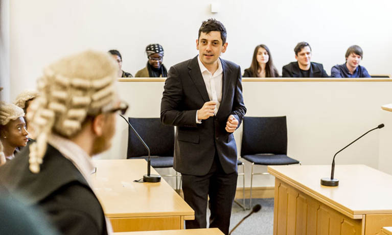 students in a mock law room - five students sitting as a jury a male standing addressing a person wearing a judges wig 