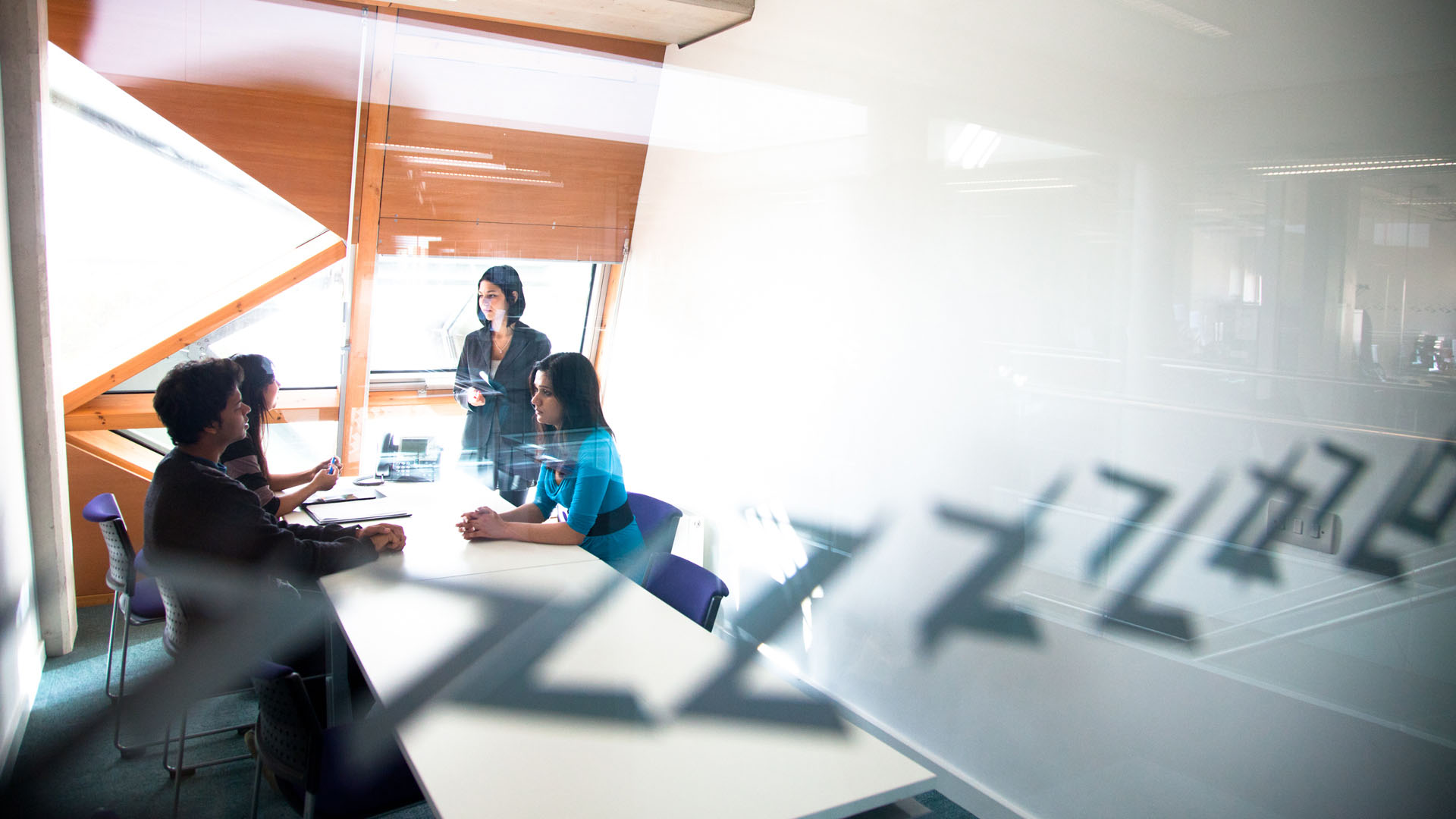 four smartly dressed people in front of a bright window face each other across a desk, one is standing