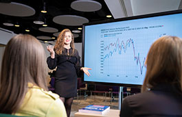 Woman standing in front of large screen, talking, while two people look at her