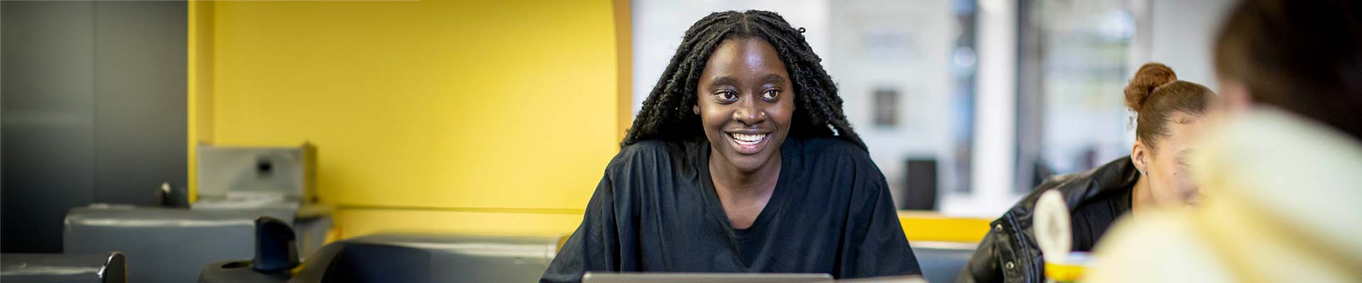 Student sitting at a table smiling at a colleague who is looking at a laptop