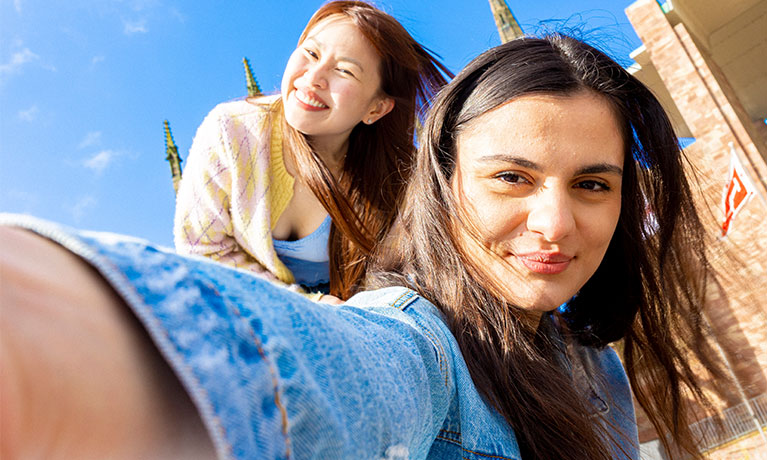 A student taking a selfie with a friend against a blue sky.