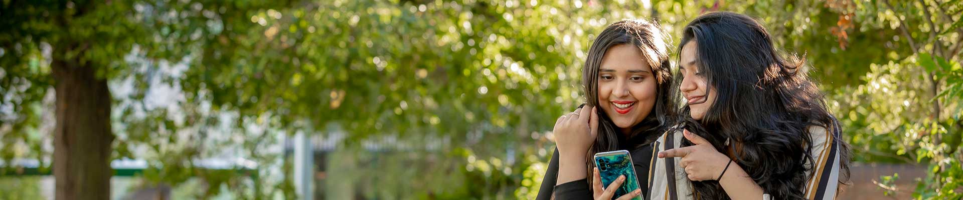 Two students sat outside of the hub looking at a phone surrounded by trees