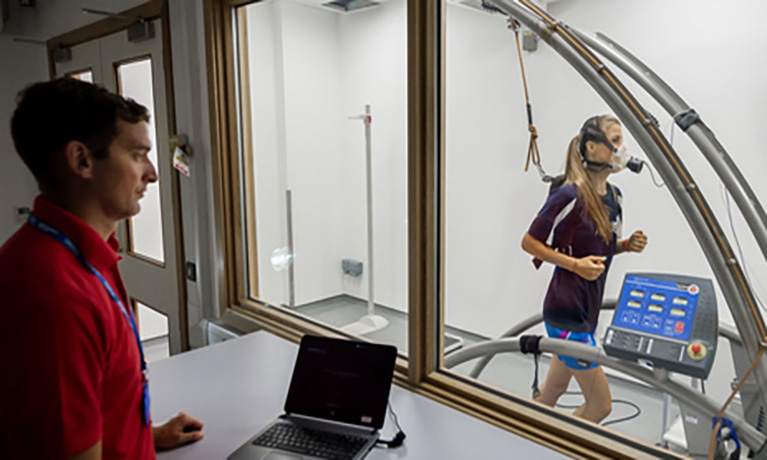 Man on laptop looking through the window at a woman on a treadmill with wires connected to her 