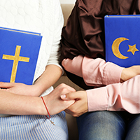 Two females sitting side by side and holding hands. One is holding a Christian Bible and the other a copy of the Quran 