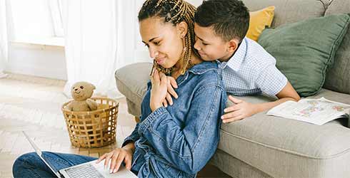 mixed race lady working on her laptop with her son hugging her neck