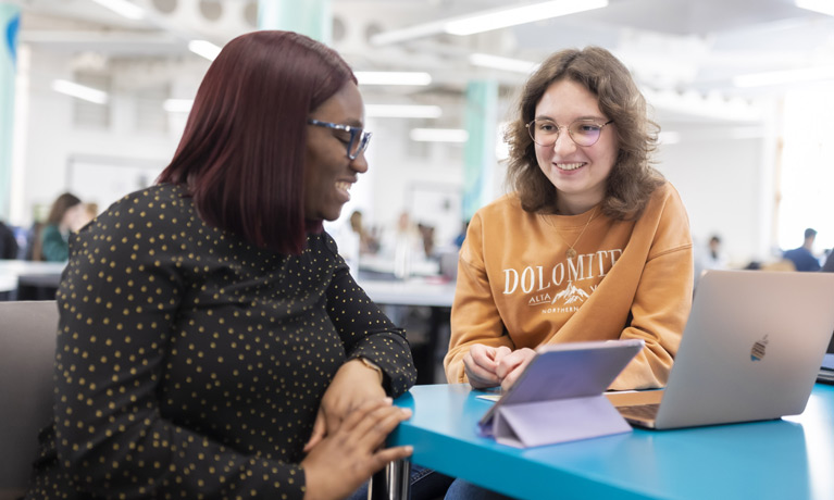 Two students in an open study area with a tablet and laptop on the table in front of them