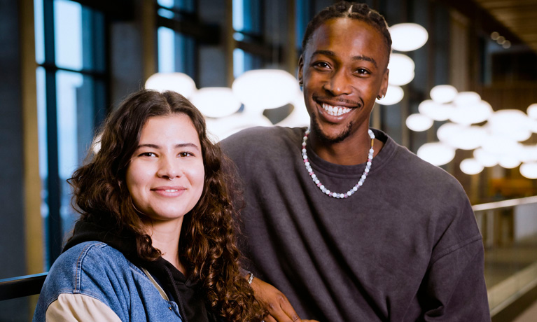 Two young, fashionably dressed students smiling at the camera with modern hanging lights in the background