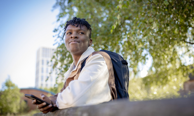 A smiling student with a blue sky and trees behind them in the background.