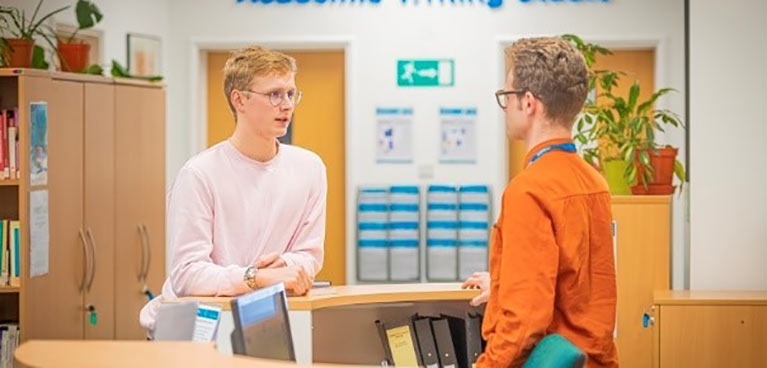 Academic support staff assisting a student at a reception desk.