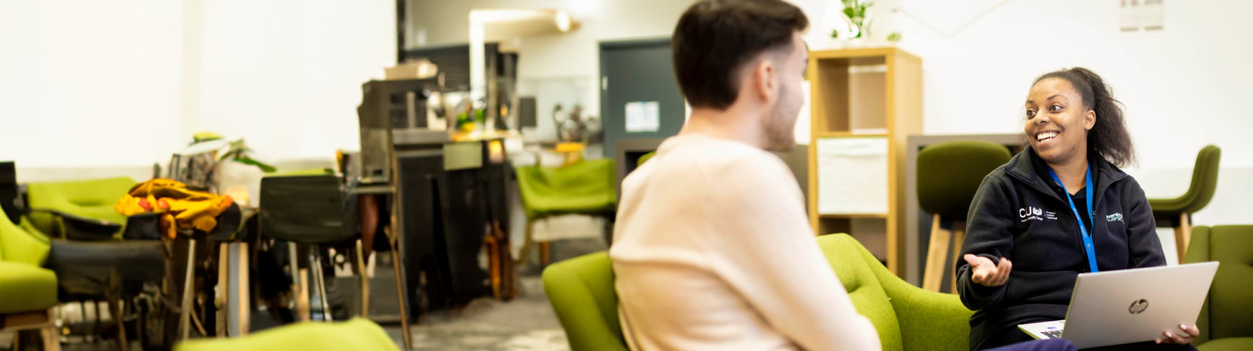 Two people sit on a sofa, engaged in conversation while one of them uses a laptop.