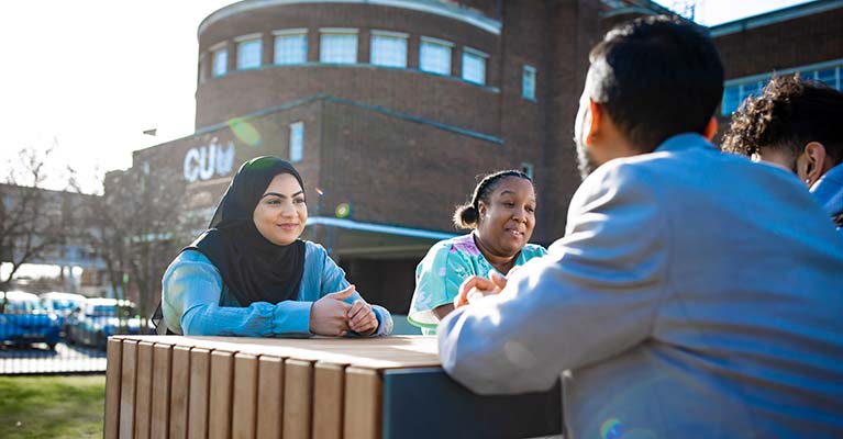 A group of students sat outside on a bench on a sunny day talking