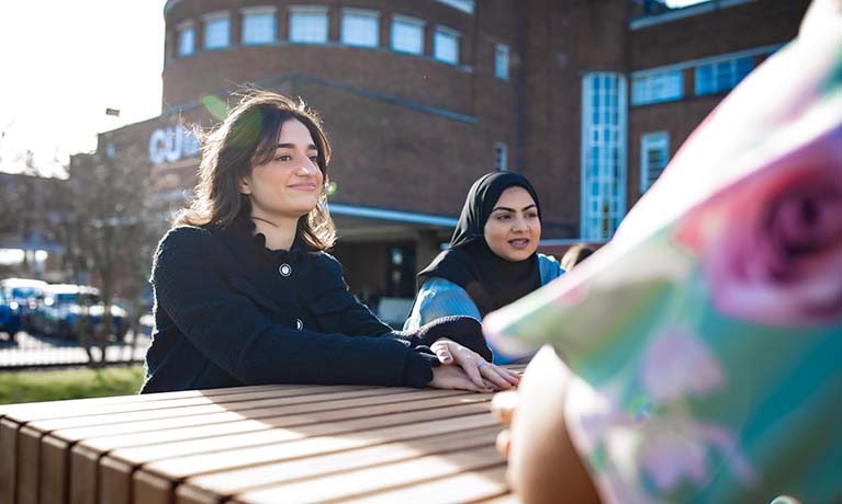 Two students sat outside on a sunny day sat on a bench talking