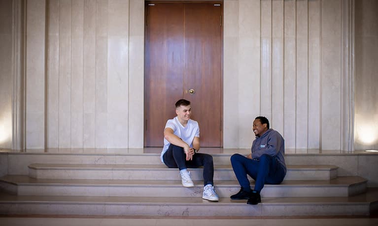 Two male students sat on a few steps inside of a building