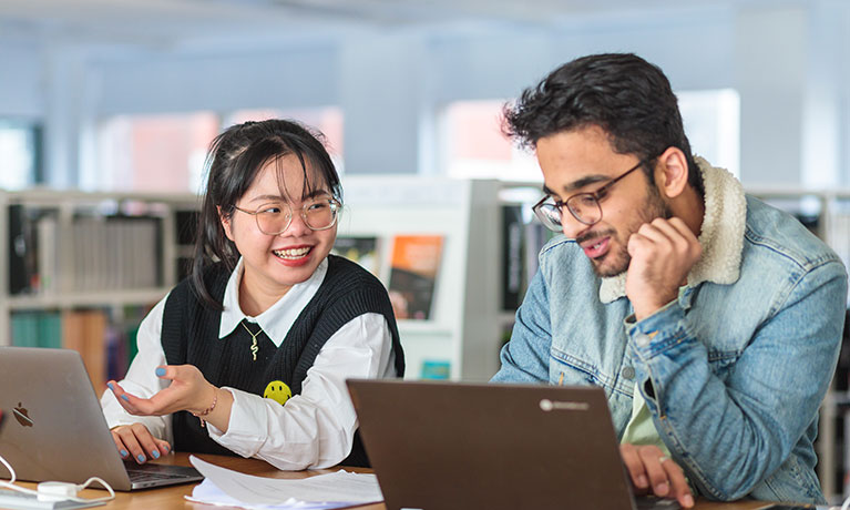 Two students sat at a desk working on their laptops