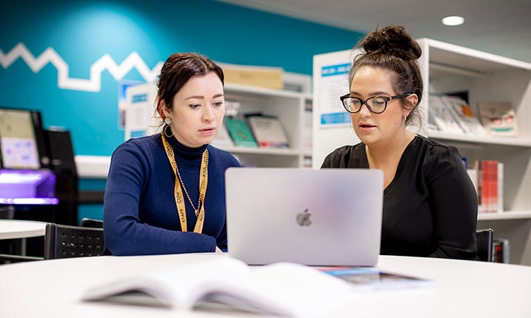 Two female students sat at a table looking at one laptop