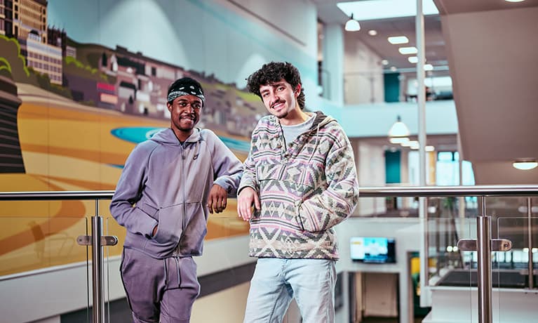 Two male students stood leaning against a glass balcony inside a building