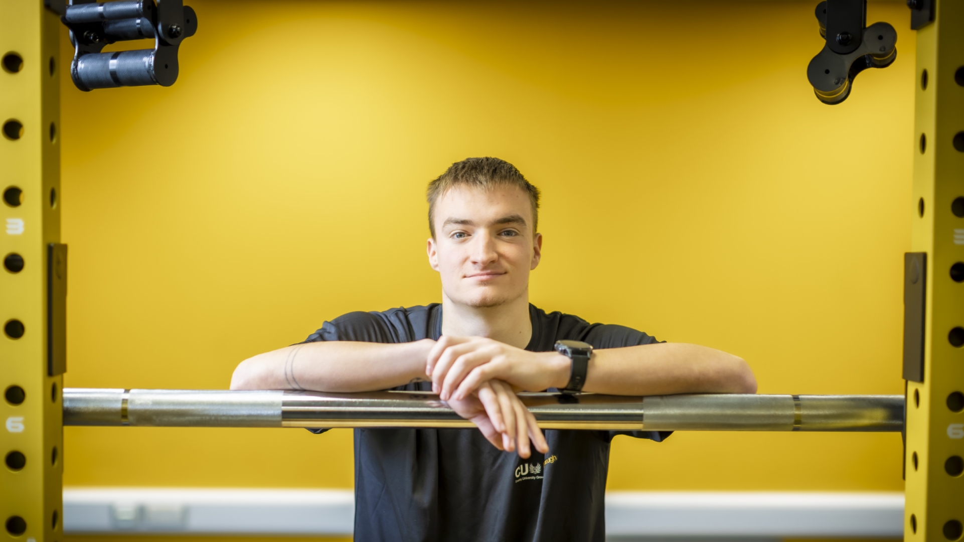 A student leaning over a weight lifting bar in a gym.