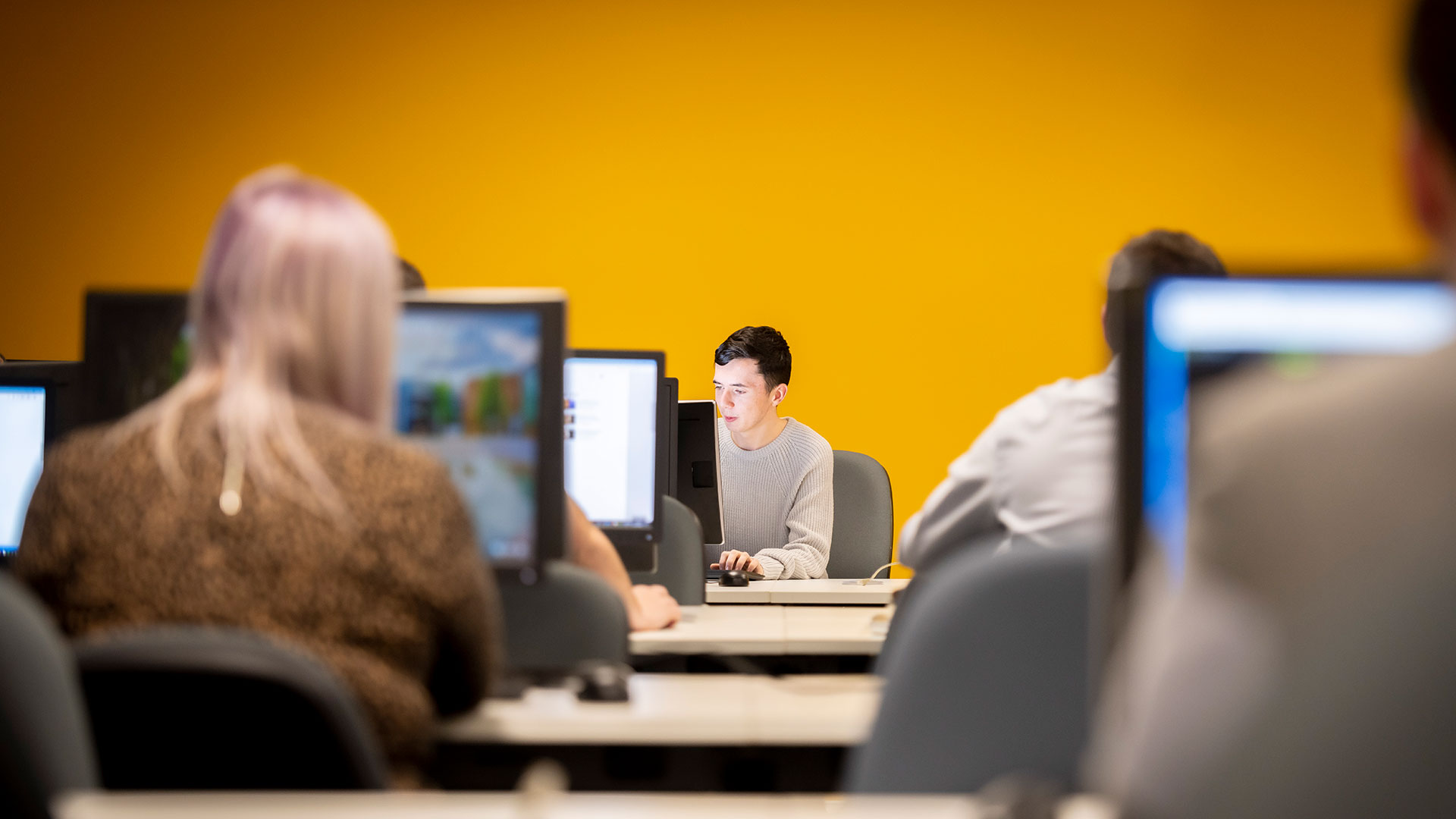 Rows of students working at banks of computers, focus is on a single student in the centre studying his screen.