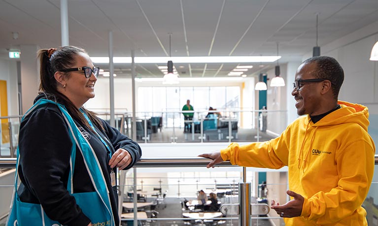 Two students talking to each other leaning against an indoor balcony