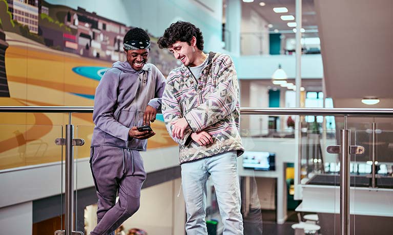 Two students leaning against a bannister indoors looking at a phone