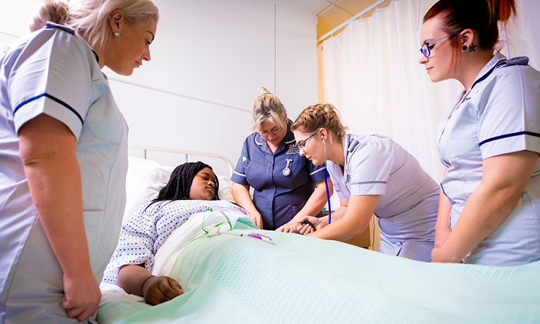 Nursing students around a mock hospital bed with a tutor and an acting student playing the role of a patient.