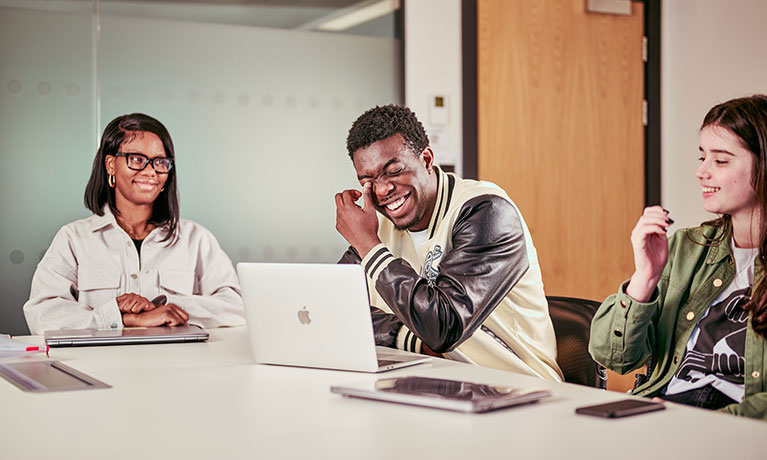 Three students sat at a desk with an open laptop laughing