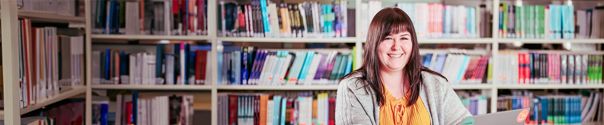 Female student sat in a library in front of a shelf of books whilst working on a laptop