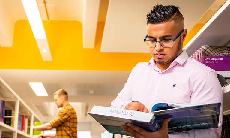 Male student stood in a library looking at a book