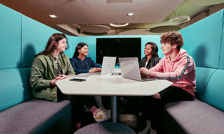 Four students sat in a pod talking with open laptops