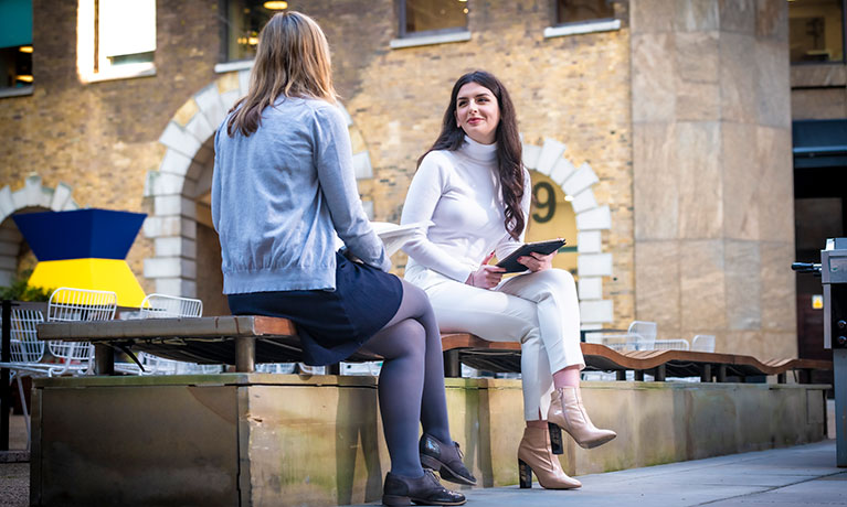 Two students sit chatting outside the London campus building