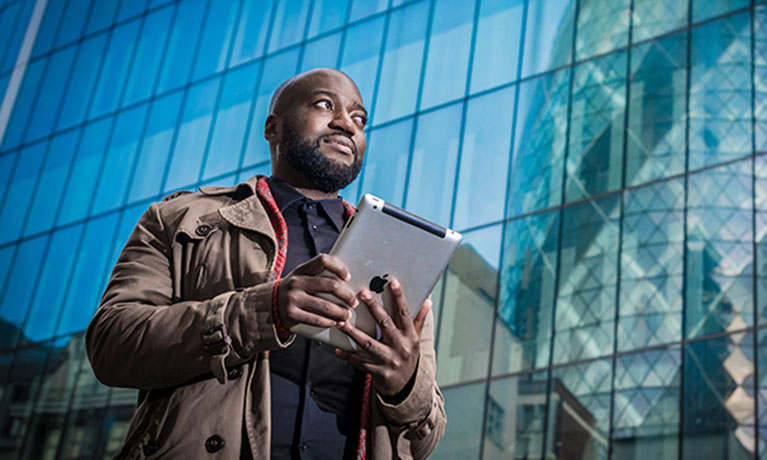 Male student stood outside of a tall building with a tablet