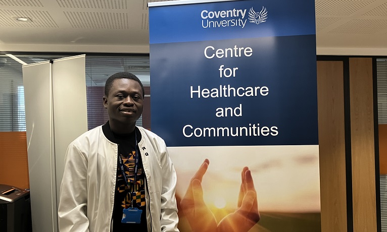 Ebenezer Akore Yeboah wearing a white jacket stood next to a banner for Coventry University's Research Centre for Healthecare and Communities