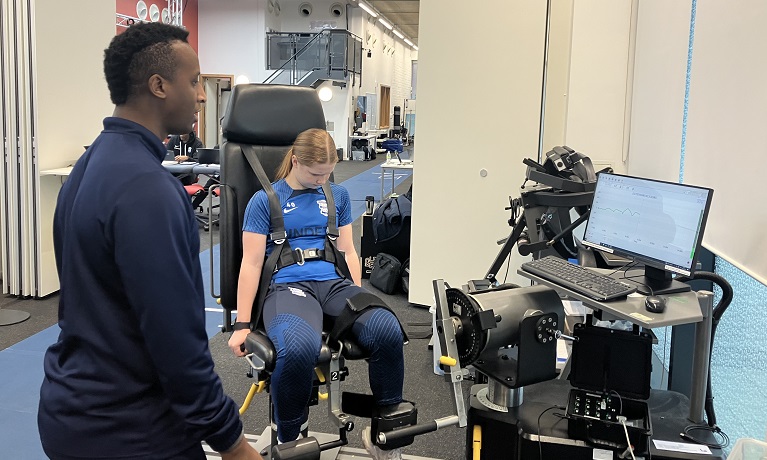 Someone sat in a chair to test their strenght with a staff member helping beside the equipment