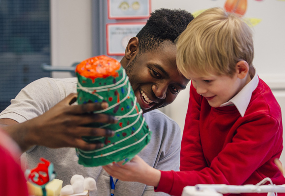 A teacher making a papier mache dinosaur with a child in the classroom.