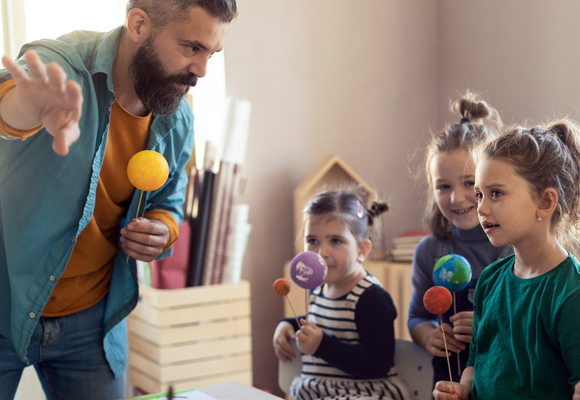 A teacher teaching children about the solar system as they hold handmade planets on sticks.