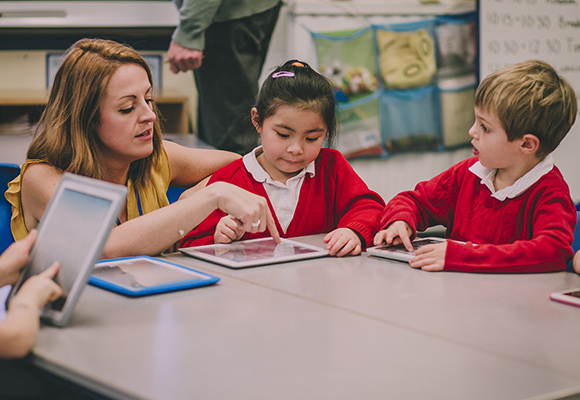 A teacher kneeling down at a table with primary age students helping them use a tablet.