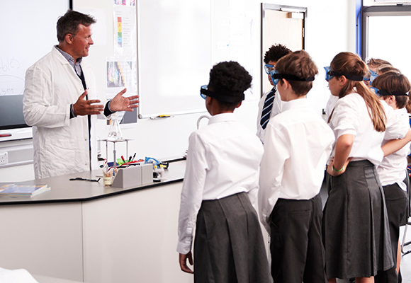 A teacher in a white coat and science lab stood in front of beackers and flasks with schoolchildren in front of him.