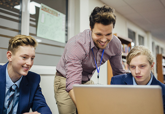 A teacher in a checked shirt helping two secondary students on a laptop.