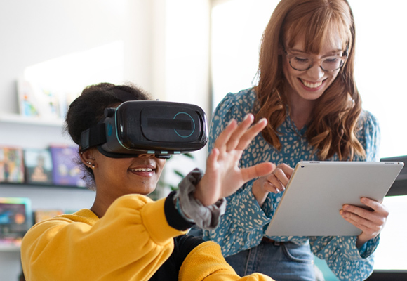 A young teacher holding a tablet while a student uses a virtual reality headset.