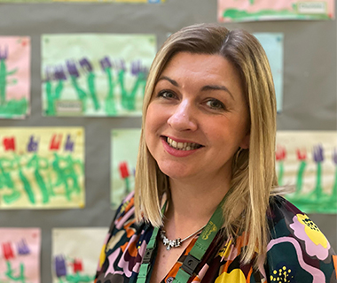 Rachel Brittain in a classroom with her schoolchildrens' paintings on the wall behind her.