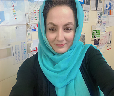 A headshot of Shagufta smiling in a classroom, with notes and diagrams on the classroom wall behind her.