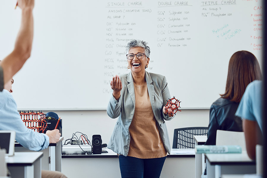 A teacher with short grey hair holding a dna helix model and laughing as students sit with their hands up.