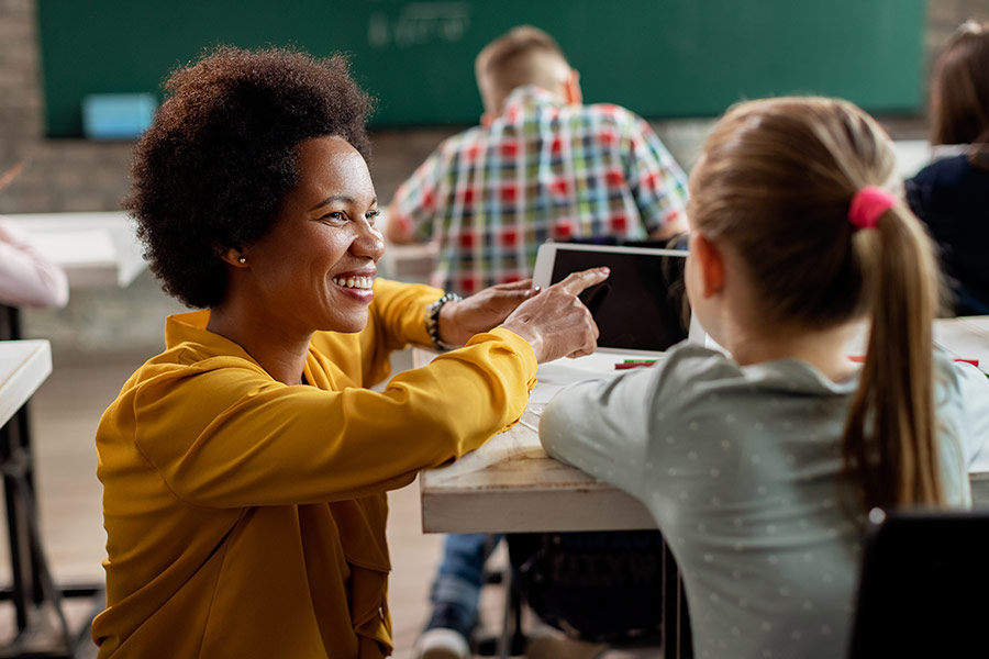 A teacher kneeling down showing a young student how to use a laptop.