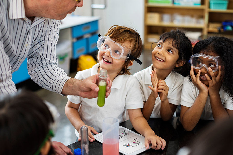 A teacher in a shirt showing three young students a test tube of green liquid.