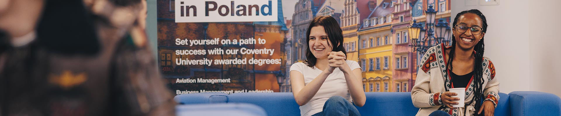 Two students sat in front of a poster on a sofa talking