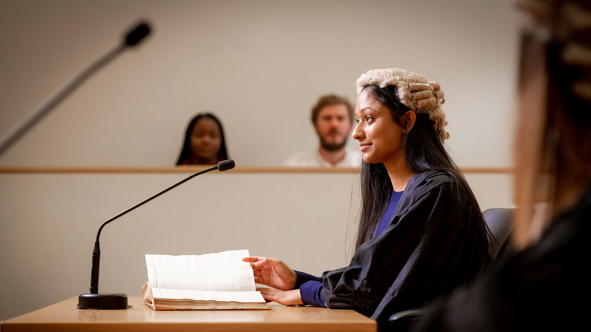 Female law student wearing robes and wig sitting in a court with notes in front of her, two people sitting behind her, blurred